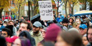 Image of a crowd of people. One is holding a sign reading "Let's stand together and Let's Love Not Hate"
