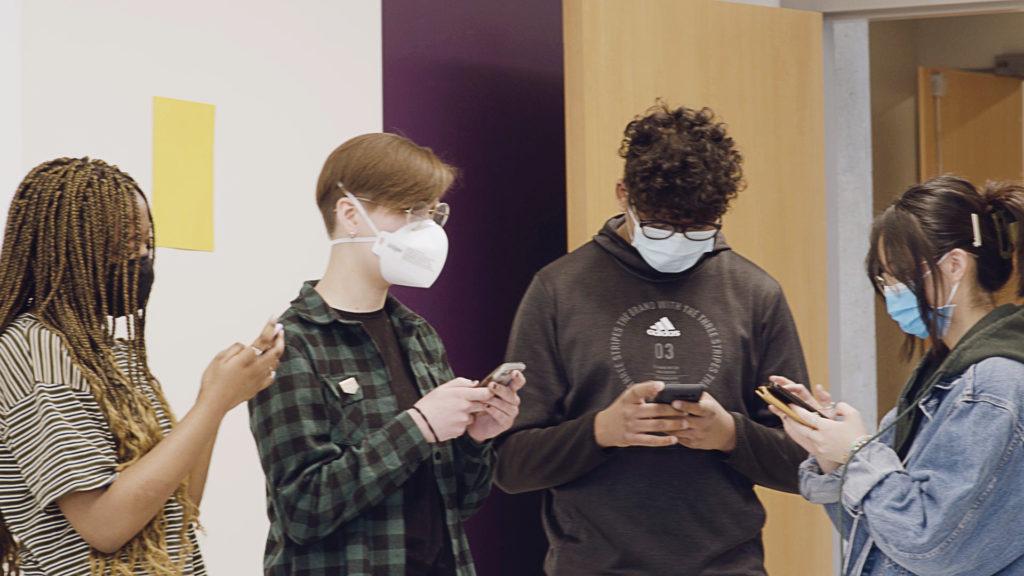 Four youth wearing masks stand in a group checking their phones as part of a Don't Click online hate and extremism prevention workshop