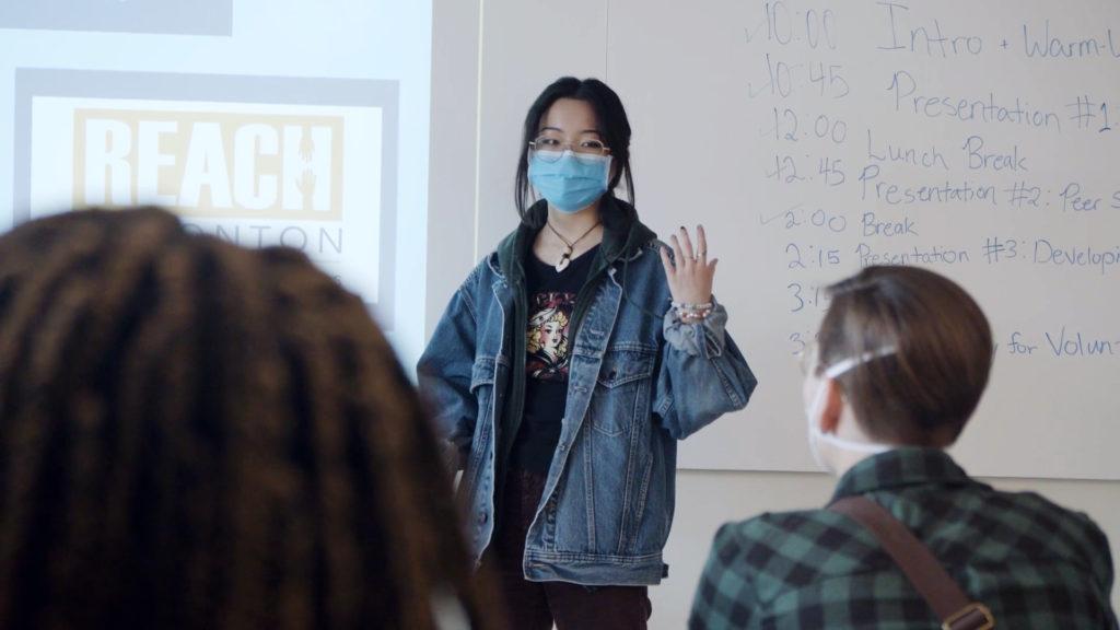 A facilitator wearing glasses, a mask, and jean jacket stands in front of a class next to a powerpoint displaying the REACH Edmonton logo as part of a Don't Click online hate and extremism prevention presentation